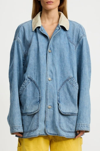 A person wears the Denimist OVERSIZED HUNTING DENIM JACKET, featuring a beige collar and large front pockets, paired with yellow pants, standing against a plain white background.