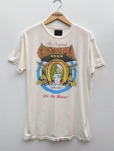 A vintage white T-shirt from Wildcat Retro Brands hangs on a wooden hanger against a plain wall. This unisex tee is adorned with a vibrant graphic featuring the words “The Original Olympia Beer” and “It’s the Water,” along with an emblem showcasing hops and a waterfall, proudly made in the USA.