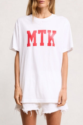 A woman in a DENIMIST CLASSIC TSHIRT by Denimist with bold red "MTK" letters and white frayed denim shorts exudes a sunfaded vibe against a light background.