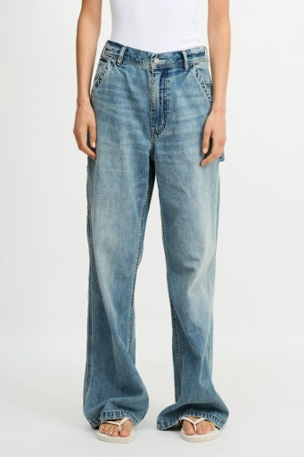 A person in loose-fitting, light blue Denimist TERI WIDE LEG CARPENTER PANT and a white sleeveless top stands with hands by their sides. The mid-rise pants reach below the ankles, partially covering white sandals.