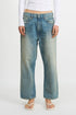Model wearing Denimist’s HAISLEY SLOUCH BF relaxed, faded blue low-rise jeans, paired with a white long-sleeve top and white flip-flops, standing against a plain white background.