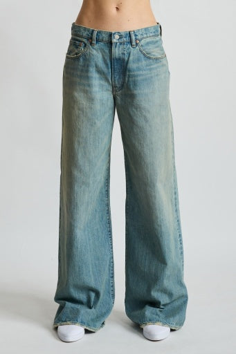 A person wears Denimist's MYLA FLARE JEAN in a relaxed, light blue faded wash with white sneakers, standing against a plain white background. The jeans have an effortless style, subtly flared and pooling at the ankles.