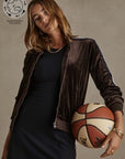 A person with shoulder-length hair in a casual outfit poses with a basketball. They wear a black dress and the brown CASTILLON KASIA JACKET by The Upside, made from recycled polyester velvet. An "Arrlow" logo and "Sustainably Focused" emblem are visible at the top left corner of the image, highlighting an eco-friendly emphasis.