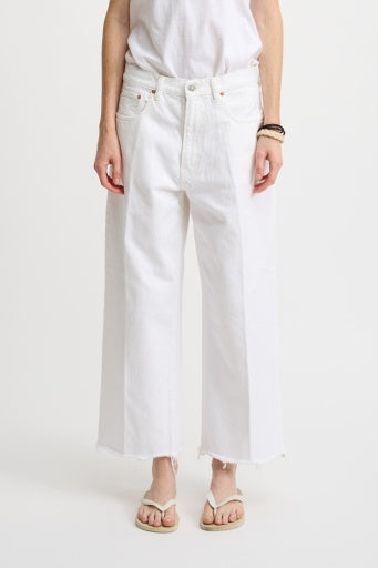 A person is wearing a white t-shirt with Denimist's GEORGIA JEAN, featuring loose-fitting, wide-legged design and frayed hems. They pair it with beige flip-flops against a plain white background.