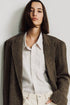 A person with straight, shoulder-length dark brown hair wears the Denimist HYANNIS DECONSTRUCTED BLAZER over a white collared shirt and cream pants, standing against a plain light background.