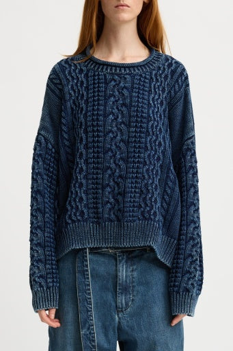 A person wearing a Denimist OVERSIZED ARAN SWEATER W/ROLLED NECK in dark blue, featuring intricate patterns and long sleeves, stands against a plain background. They are paired with loose-fitting jeans, but their face is obscured.