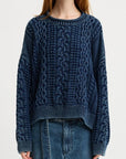 A person wearing a Denimist OVERSIZED ARAN SWEATER W/ROLLED NECK in dark blue, featuring intricate patterns and long sleeves, stands against a plain background. They are paired with loose-fitting jeans, but their face is obscured.