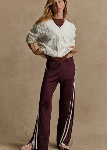 Against a neutral background, a person wears The Upside's JULIET PANT in low-rise maroon with white stripes, and a slinky fit for style. An elastic waist ensures comfort. They're sporting a white cable-knit sweater over a maroon top, with long hair and sneakers, hands casually in pockets.