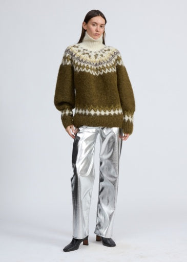 A woman with long brown hair and a neutral expression models the Rag & Bone beth oversized fair isle tneck sweater, featuring green and white patterns, paired with silver metallic pants and black shoes against a plain background.