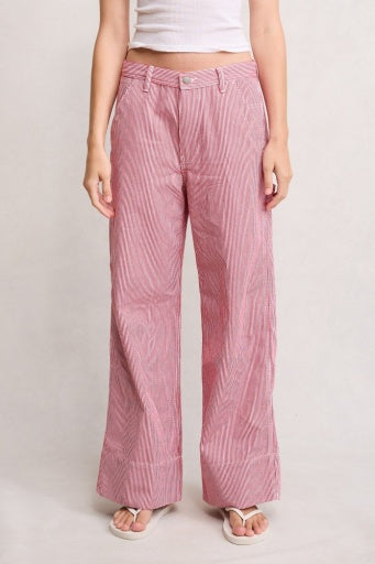 A person wears Denimist's PAYDEN RELAXED CARPENTER PANT—wide-leg, high-waisted, pink and white striped—styled with a white sleeveless crop top and white sandals, standing against a plain light background.