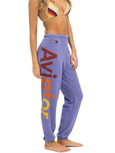 A woman is standing sideways wearing a multi-colored bikini top and purple Aviator Nation 2 - Womens Sweatpants with the word "Aviator Nation" printed in large letters down the leg. The word "Aviator Nation" is displayed vertically in red and yellow text. The background is plain white.