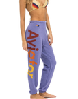 A woman is standing sideways wearing a multi-colored bikini top and purple Aviator Nation 2 - Womens Sweatpants with the word "Aviator Nation" printed in large letters down the leg. The word "Aviator Nation" is displayed vertically in red and yellow text. The background is plain white.