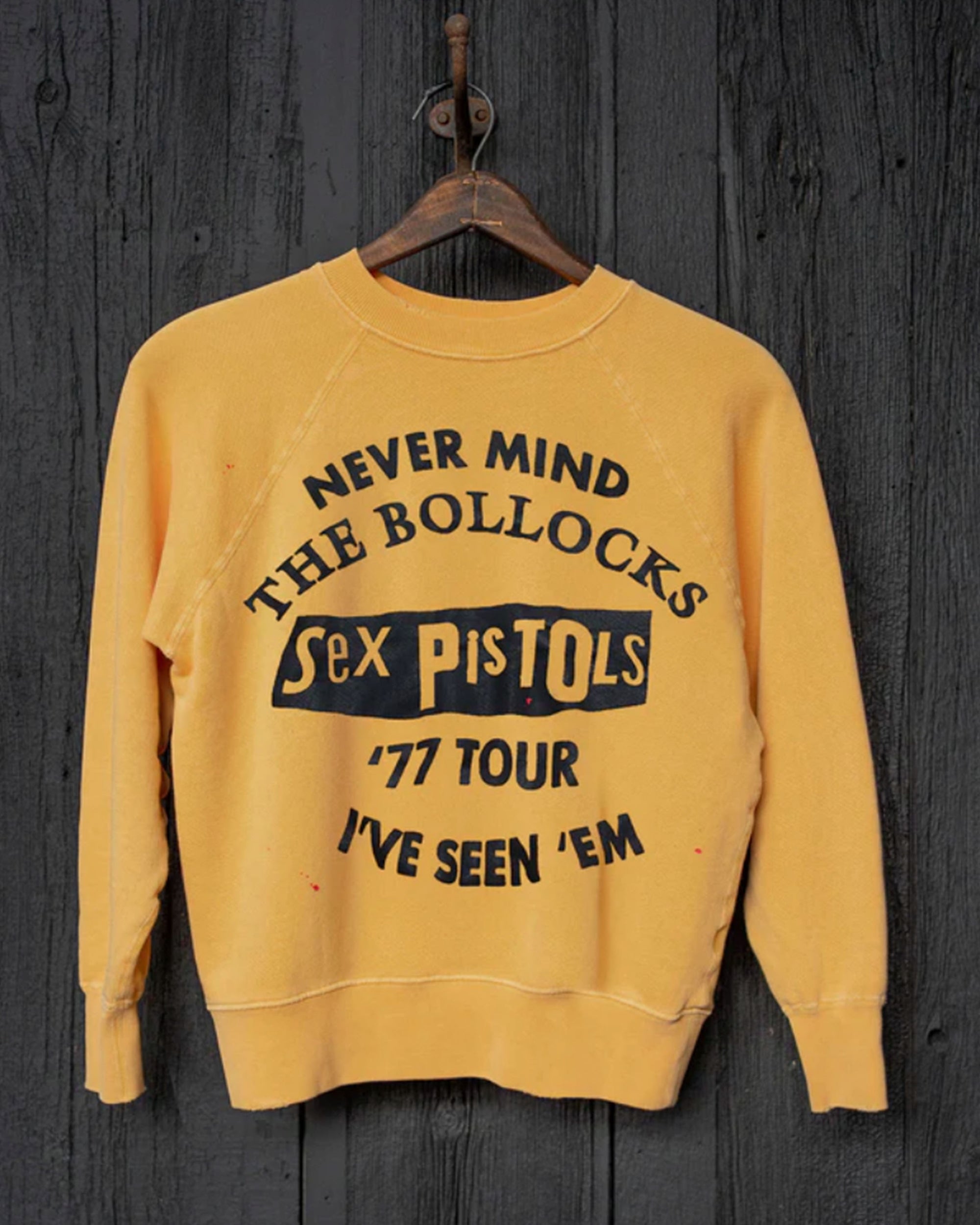 The Made Worn Sex Pistols Shrunken LS, a vintage yellow heavyweight cotton sweatshirt with black “Never Mind The Bollocks, Sex Pistols, '77 Tour, I've Seen 'Em” text, hangs on a wooden hanger against a dark wood wall. Made in USA.