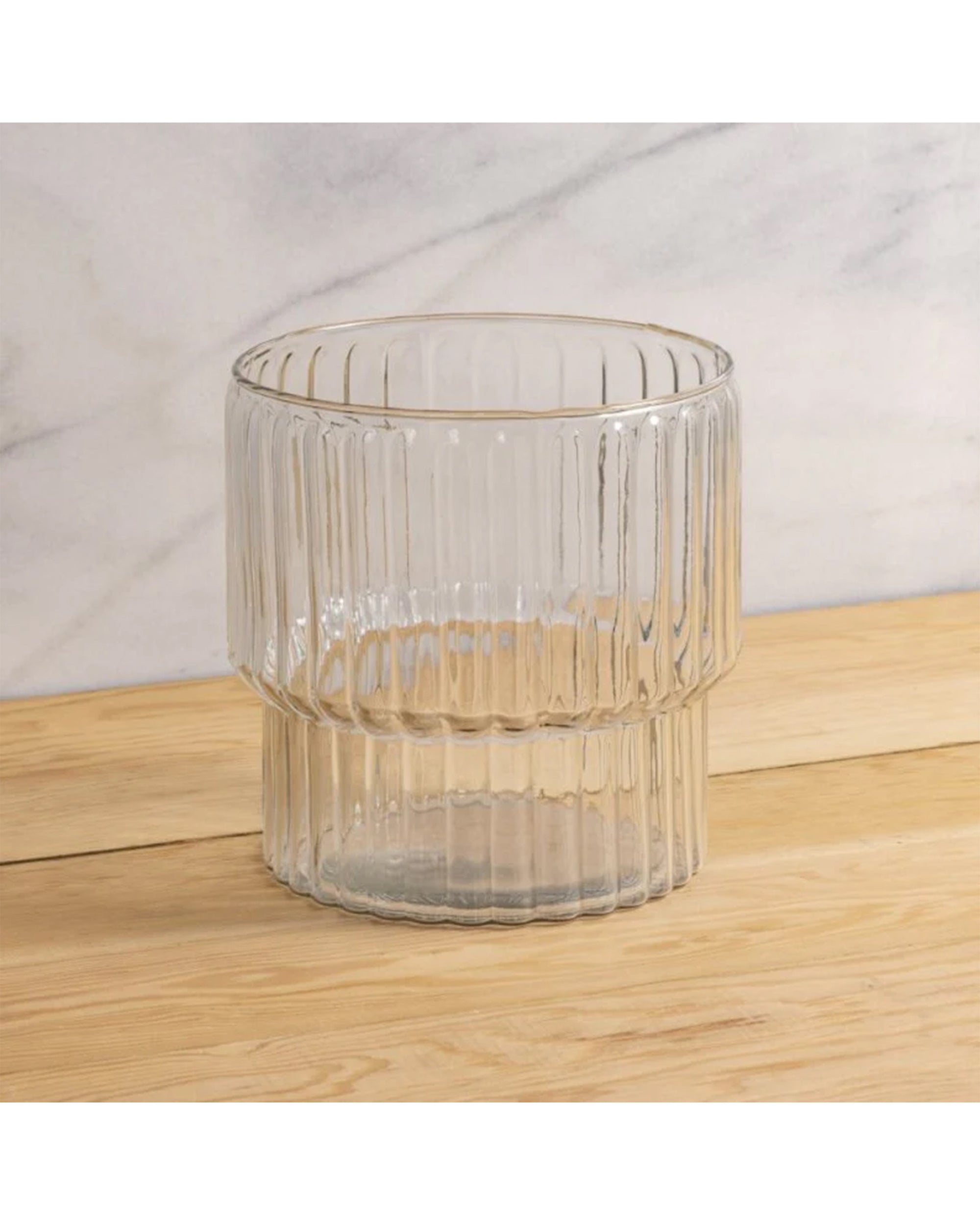 The Shae Champagne Bucket by Be Home, with its clear ribbed glass and vertical texture, rests on a light wooden surface against a white and gray marbled background—ideal for elevating any luxury entertaining setting.