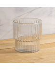 The Shae Champagne Bucket by Be Home, with its clear ribbed glass and vertical texture, rests on a light wooden surface against a white and gray marbled background—ideal for elevating any luxury entertaining setting.
