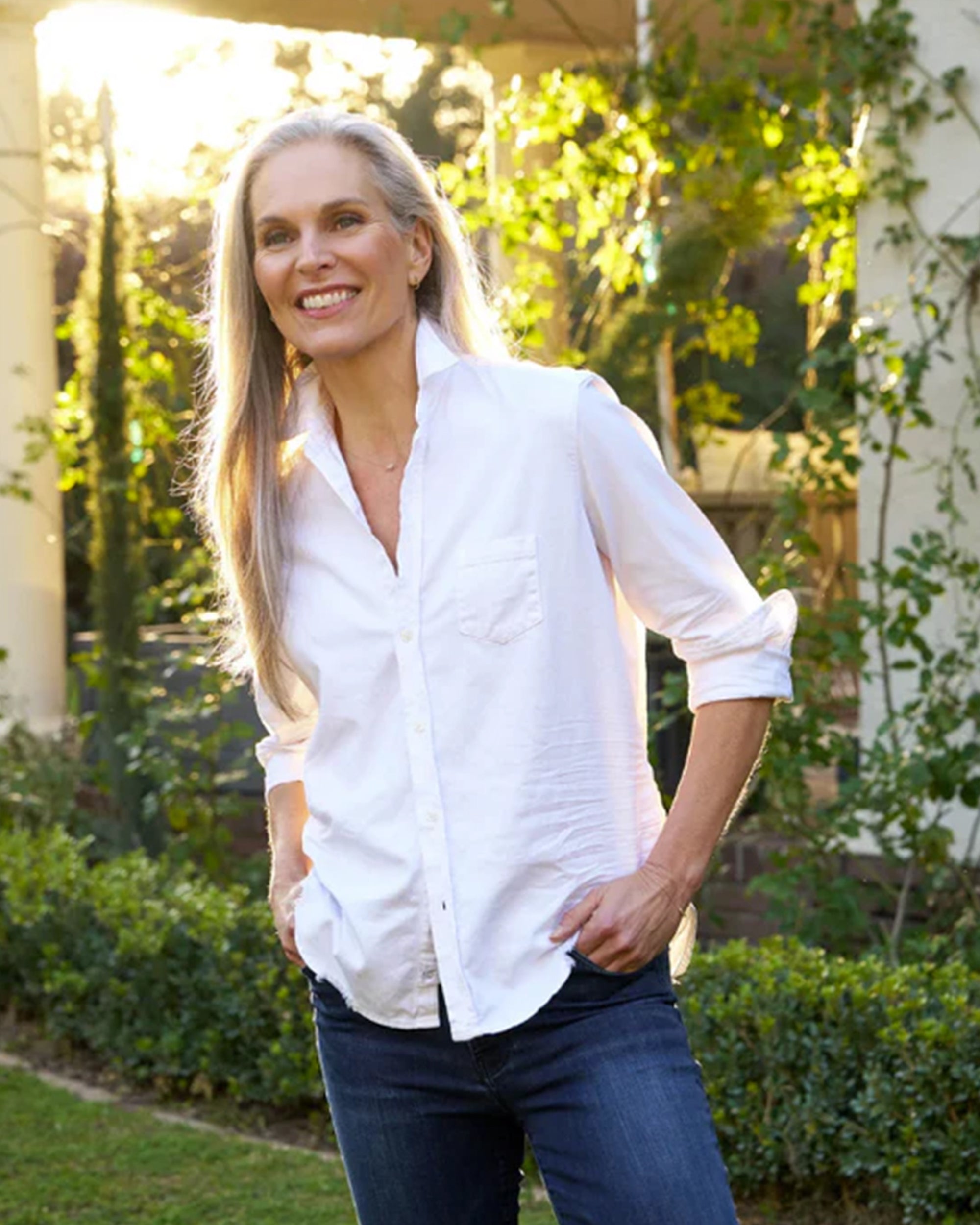 A smiling woman with long gray hair stands in a sunlit garden, wearing Frank & Eileen's Shirley Tattered Denim and a relaxed white button-up shirt, her hands in her pockets as sunlight filters through the greenery.