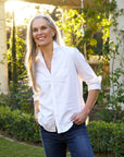 A smiling woman with long gray hair stands in a sunlit garden, wearing Frank & Eileen's Shirley Tattered Denim and a relaxed white button-up shirt, her hands in her pockets as sunlight filters through the greenery.