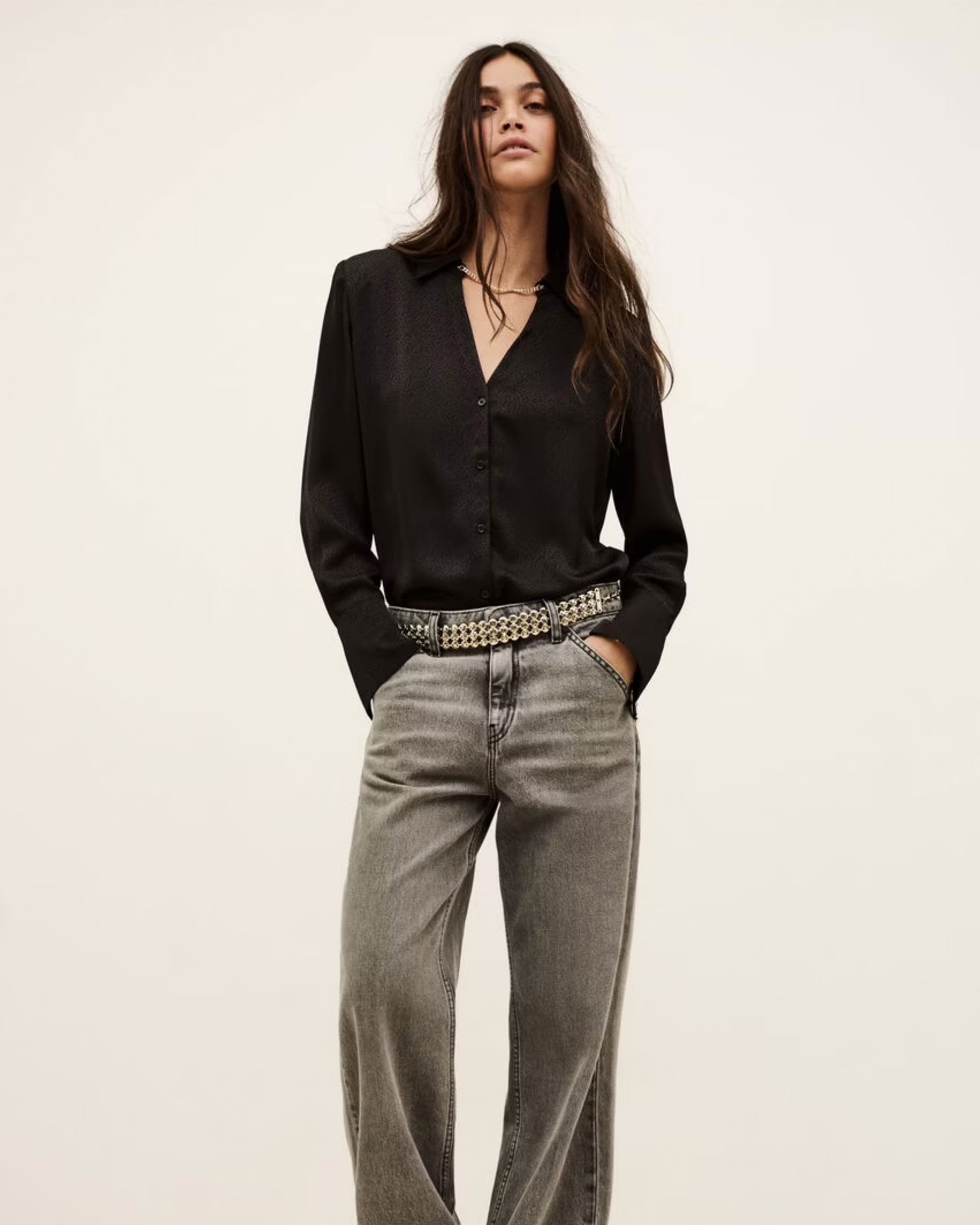 A woman with long, wavy dark hair stands against a neutral background, wearing the BA&SH Shirt Itsey— a black button-up with loose shoulders—tucked into high-waisted, loose grey jeans and styled with a silver studded belt. Hands in pockets.