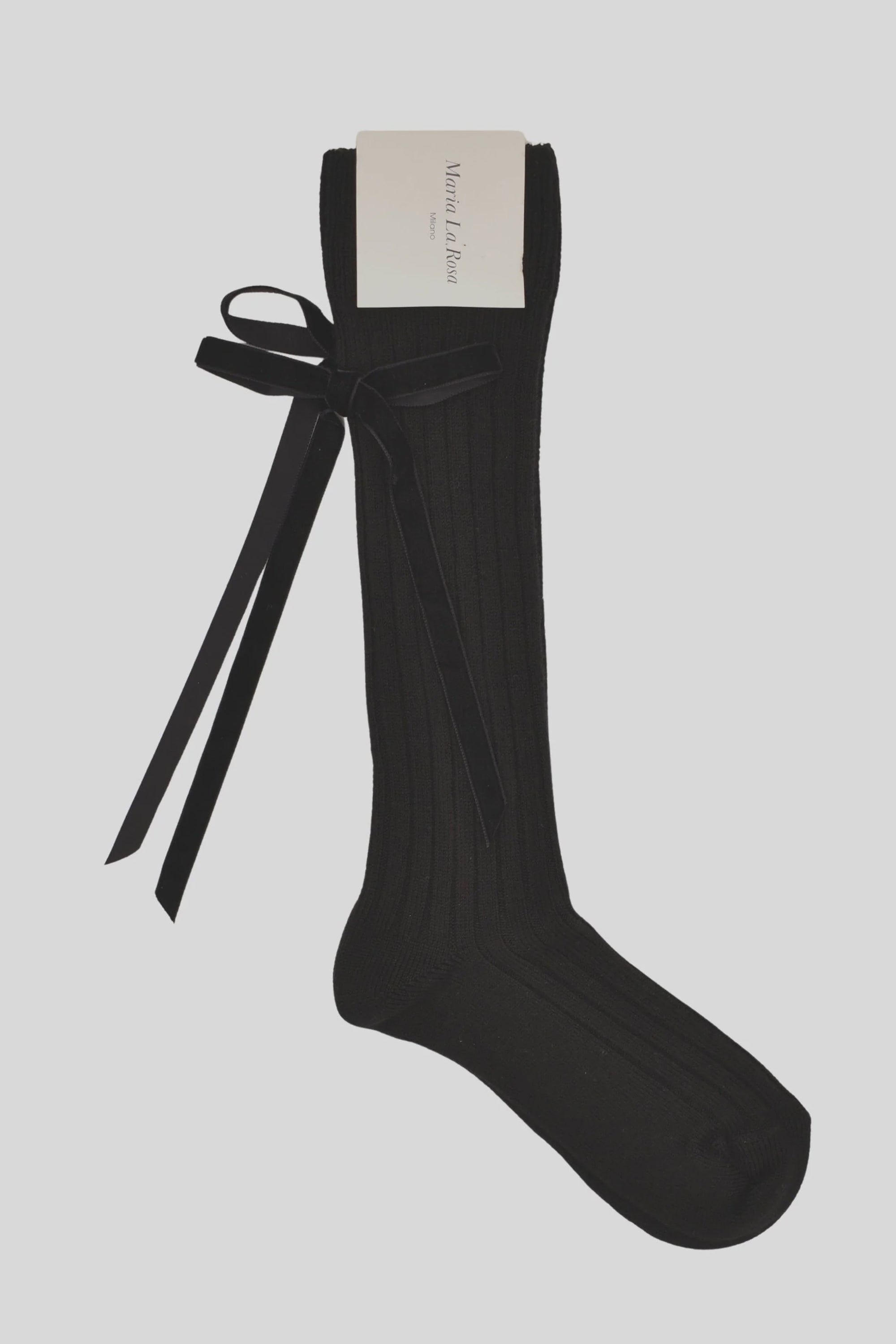 The Maria La Rosa College Velvet Bow Sock is a single black knee-high with vertical ribbing and a large velvet bow on the side, shown against a plain light background, featuring a white tag at the top.