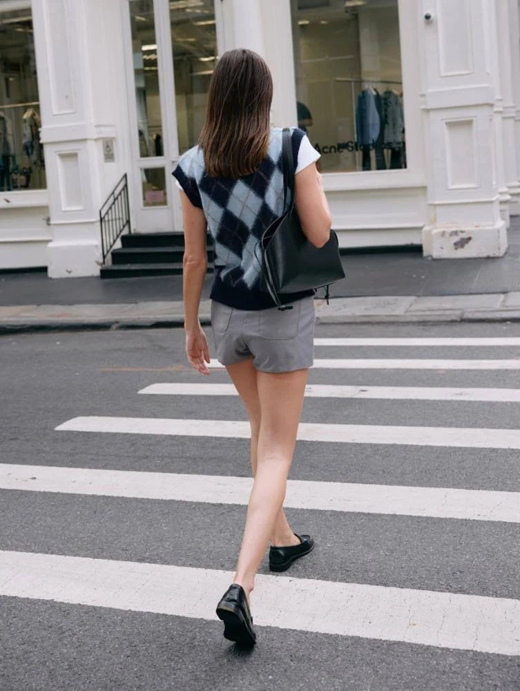 A woman with straight brown hair, in a White + Warren Mohair Luxe Argyle Vest, gray shorts, and black shoes, carries a black bag as she crosses a city street at a crosswalk.