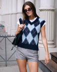 A woman wearing sunglasses, the White + Warren Mohair Luxe Argyle Vest over a white t-shirt, and gray shorts walks outdoors near white columns, carrying a black bag on her shoulder.