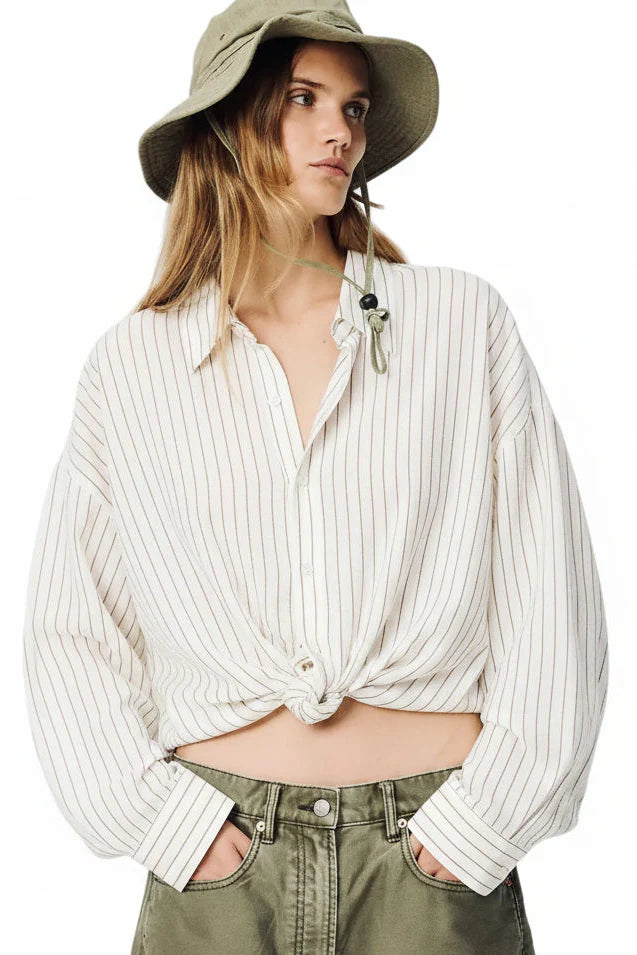 A person in the Denimist TWISTED FRONT SHIRT—a relaxed-fit linen-cotton top with thin vertical stripes and a front tie—paired with a wide-brimmed khaki hat and olive green pants stands against a white background, hands in pockets.