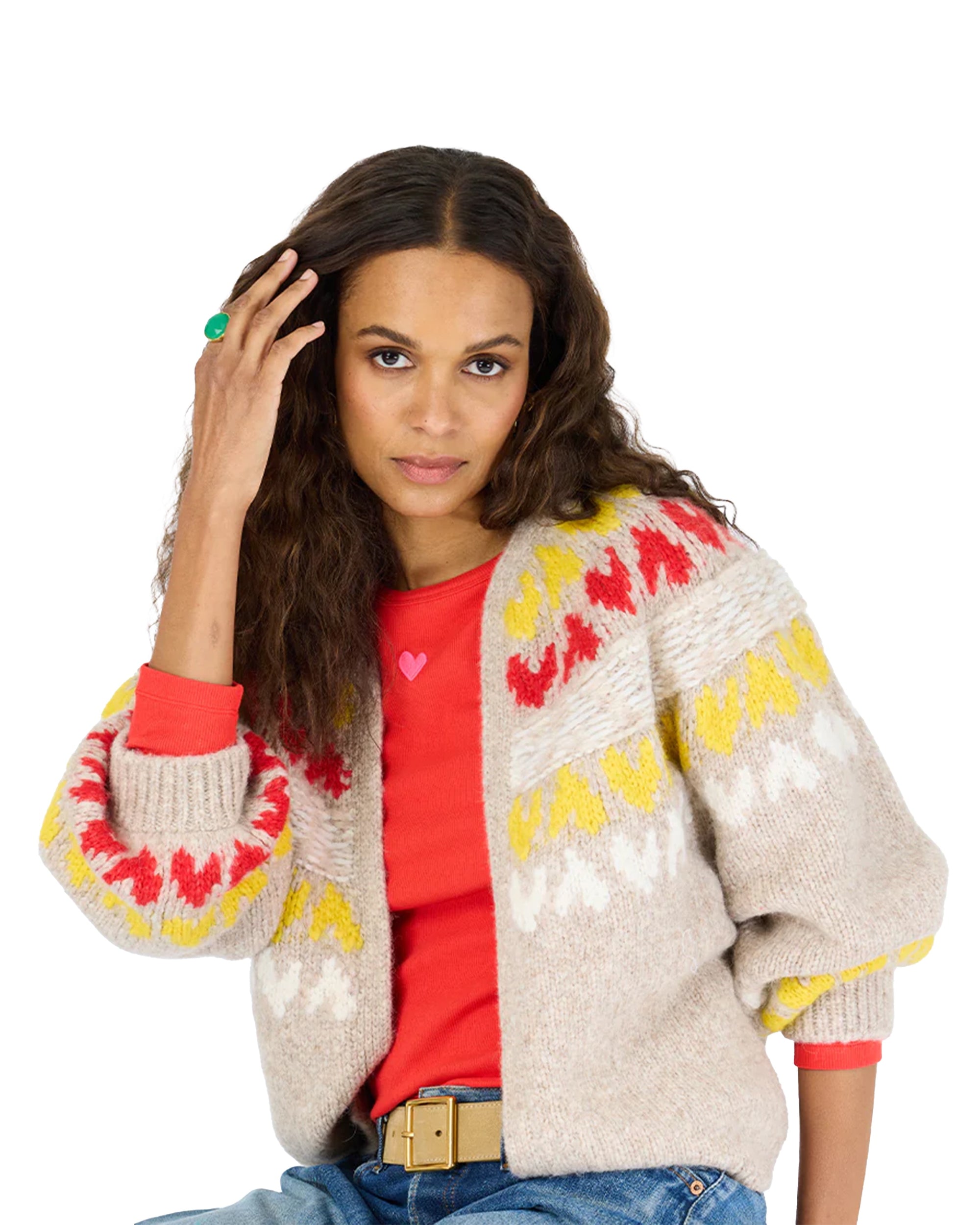 A woman with long wavy brown hair wears the Kerri Rosenthal Sienna Ker-isle Cardigan, featuring red and yellow patterns, over a red shirt with a pink heart. She sits in blue jeans and a green ring, touching her hair with one hand.