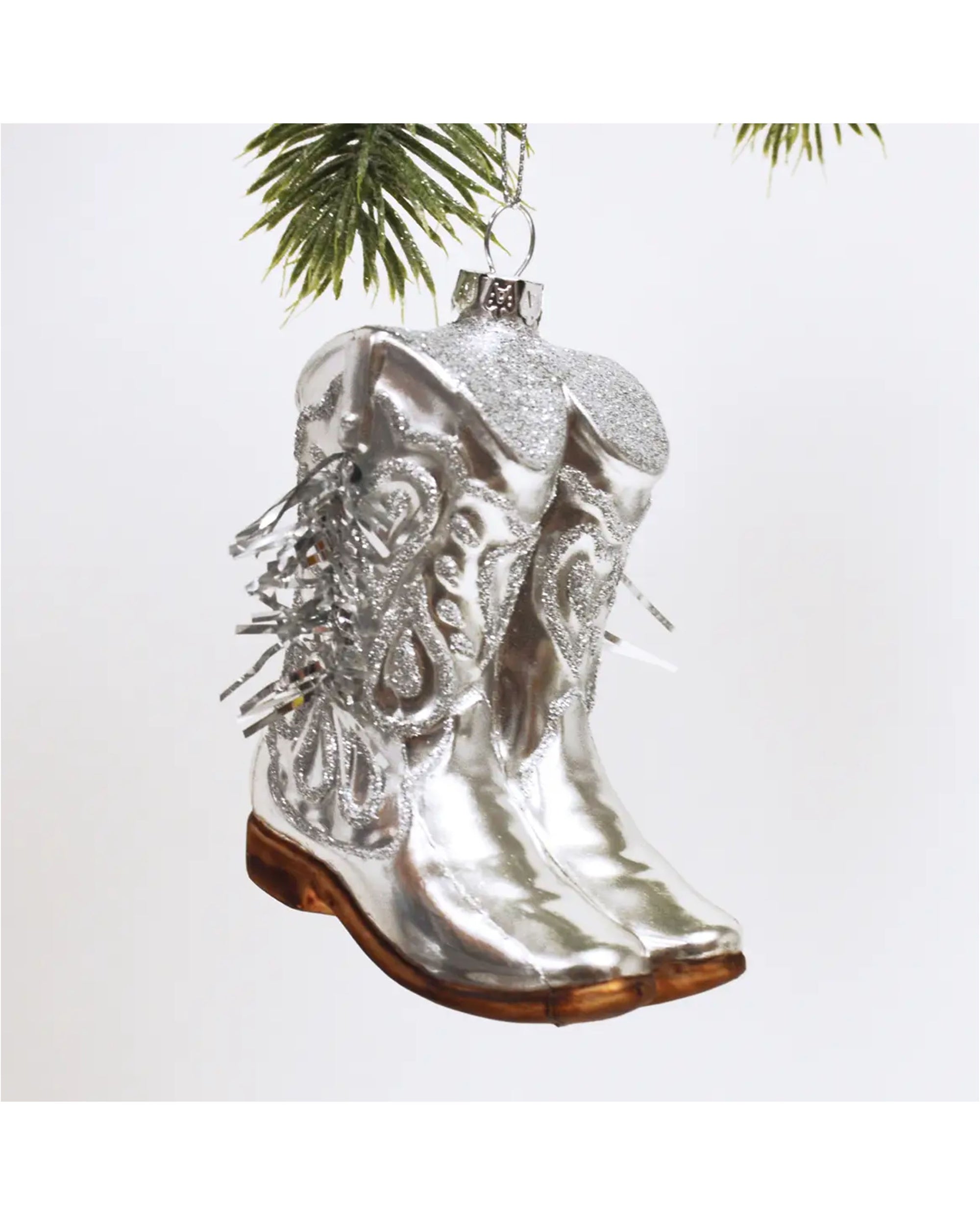 The Faire Cowgirl Boot Ornament features shiny silver with glitter details and brown soles, hanging from your Christmas tree to add a western flair to your holiday décor.