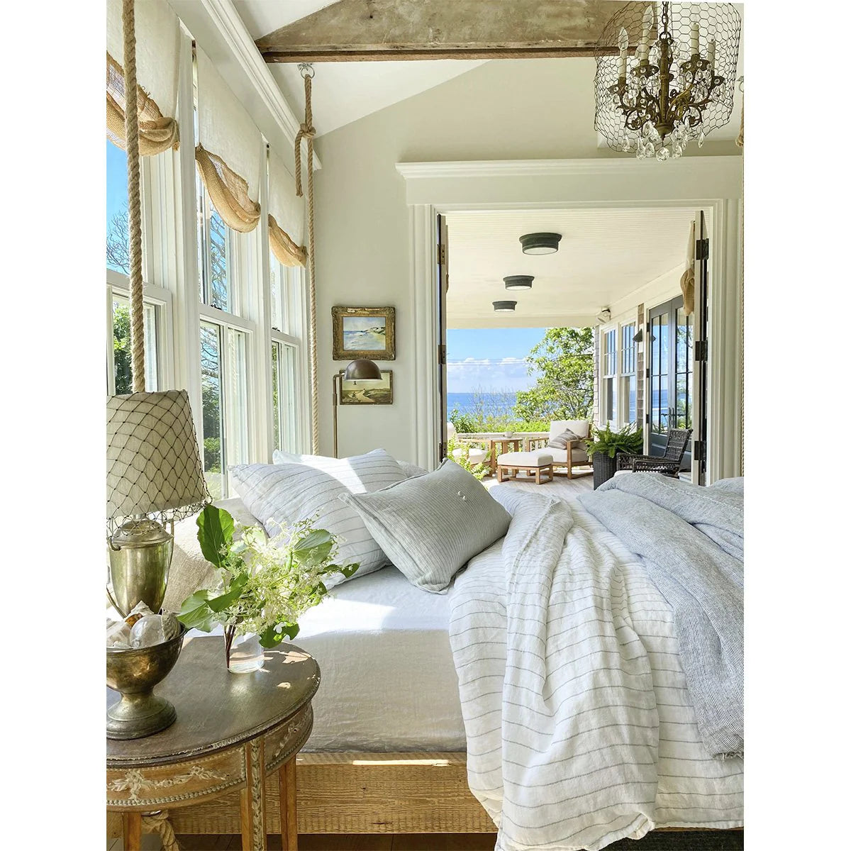 The image presents a well-lit bedroom showcasing white bedding from the Lake Linen Collection, accompanied by a bedside table featuring a lamp and vase. Through large windows, you can see views of a porch with seating arrangements and the serene blue ocean in the distance, framed by lush greenery. A chandelier hangs gracefully from the ceiling, enhancing the room&#39;s elegance with its presence next to POM POM&#39;s Blake Big Pillow 28x36.
