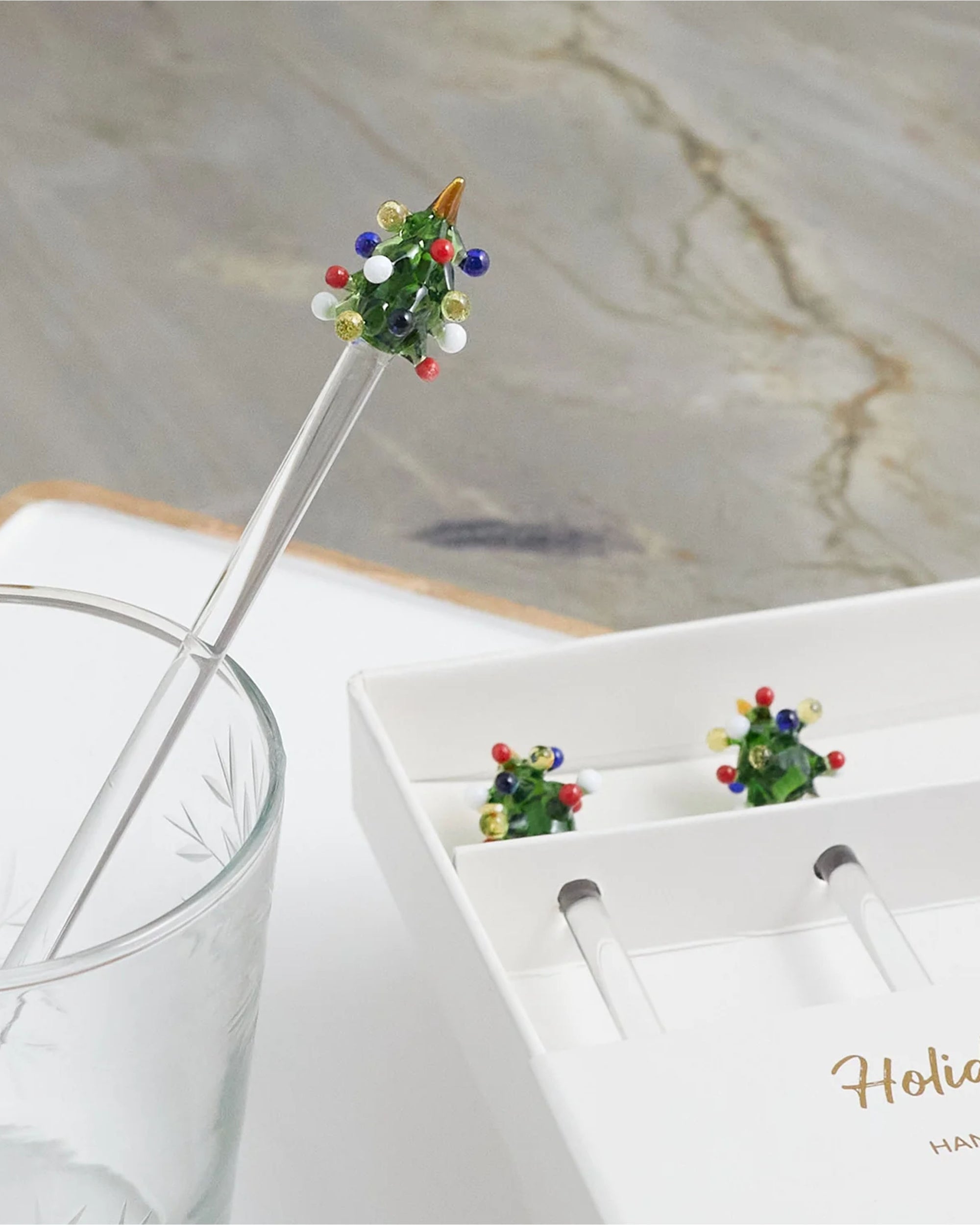 A Be Home Tree Glass Swizzle Stick, featuring a festive Christmas tree topper, sits in a glass beside a white box holding two more from the Tree Glass Swizzle Sticks S/4 set.