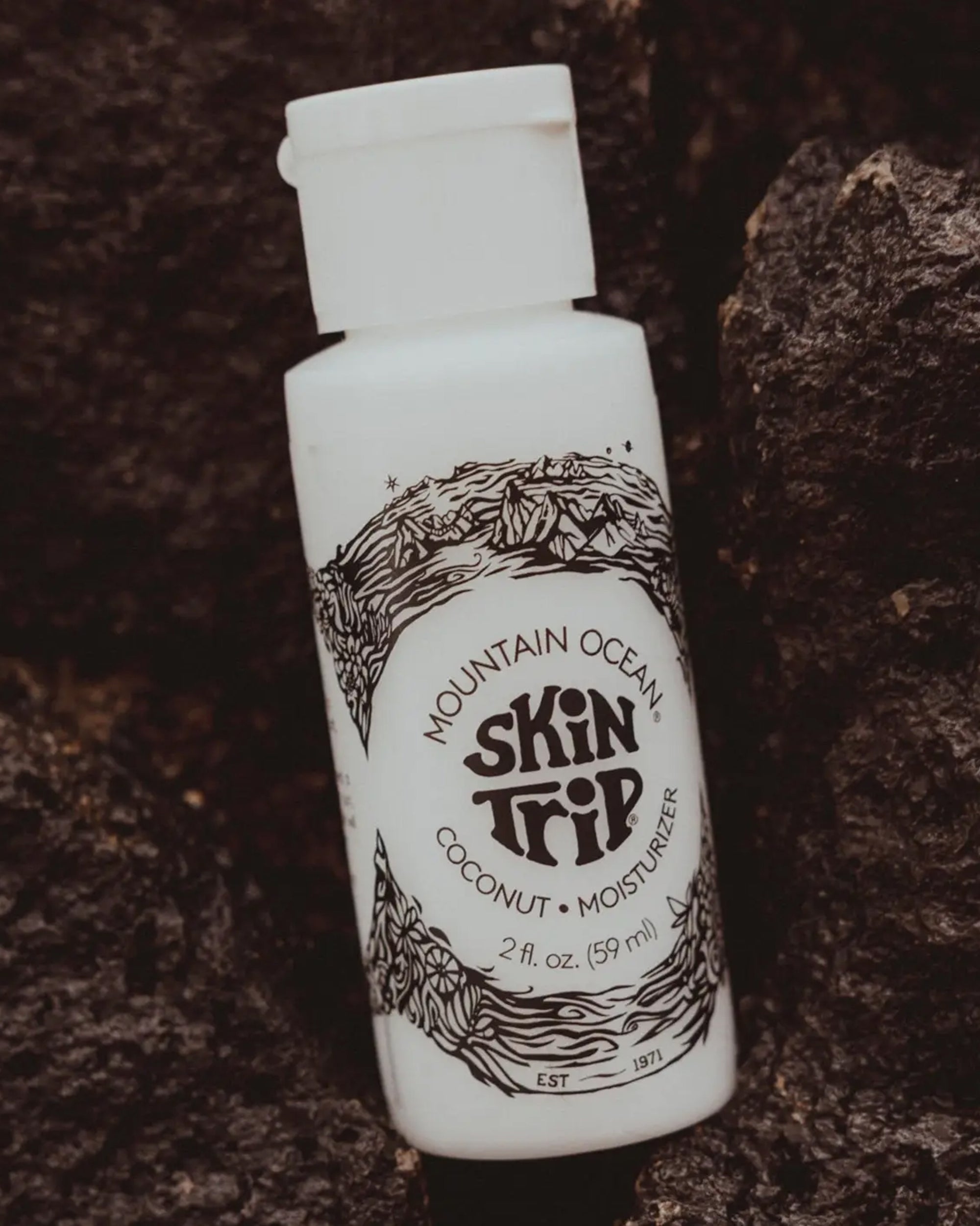 A white bottle of Faire Skin Trip Travel Size sits on dark, textured rocks. This coconut-scented lotion features black mountain and wave art, offering natural skincare in a convenient travel-size moisturizer.