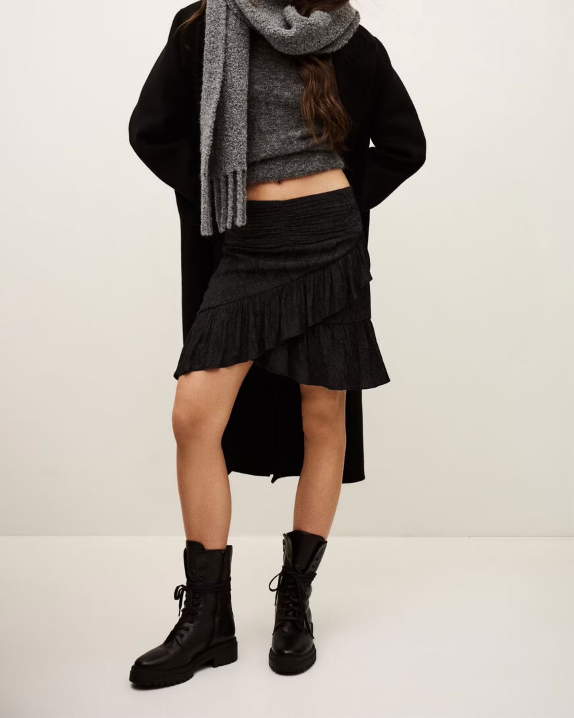 A person wears the BA&SH Skirt Rino with a grey polyester scarf, grey sweater, black coat, and black lace-up boots against a plain light background. Their face is not visible.