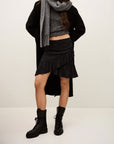 A person wears the BA&SH Skirt Rino with a grey polyester scarf, grey sweater, black coat, and black lace-up boots against a plain light background. Their face is not visible.