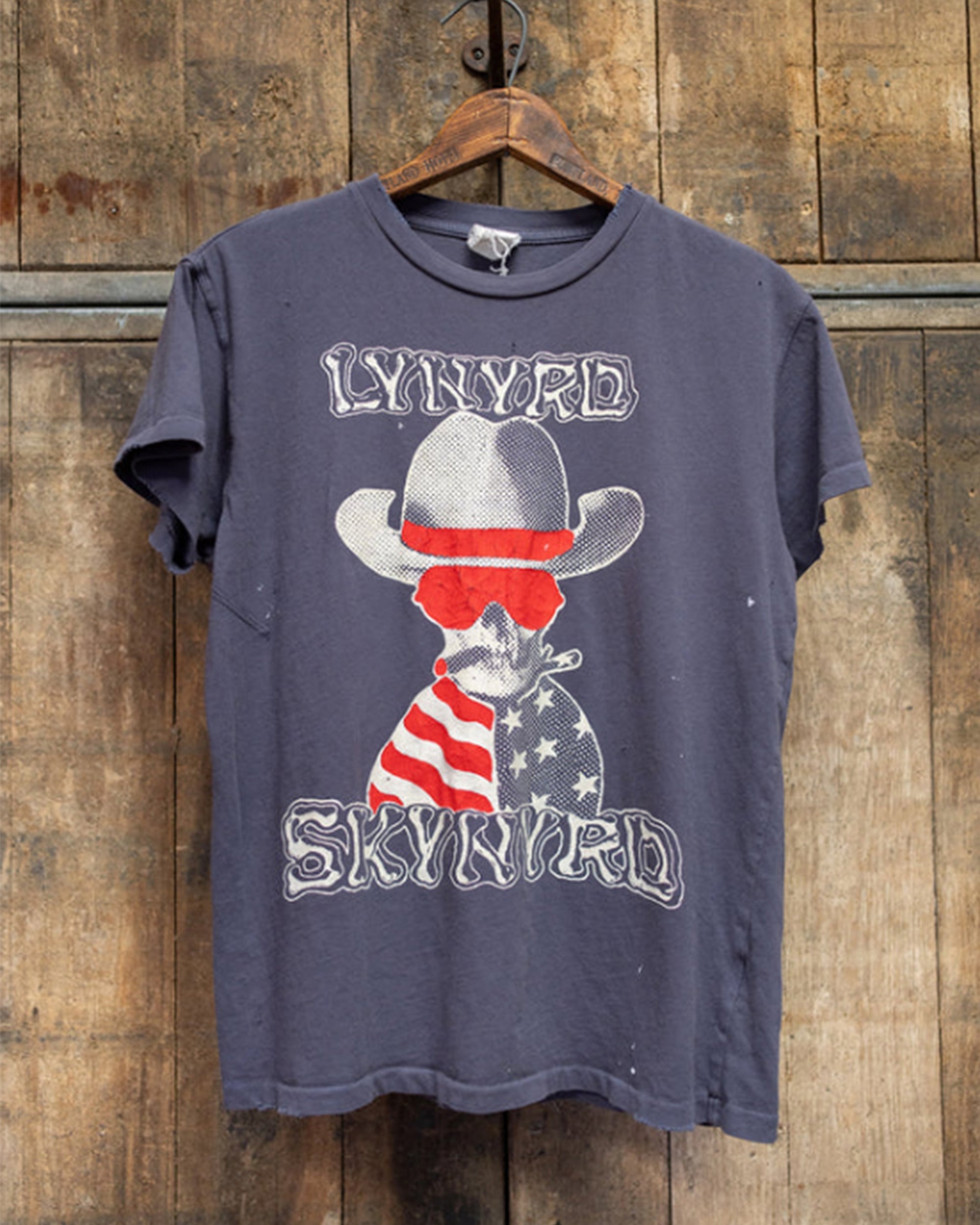 A faded vintage-style Lynyrd Skynyrd Crew Tee by Made Worn, 100% cotton and made in the USA, hangs on a wooden hanger. It features a dog in a cowboy hat with the American flag and "Lynyrd Skynyrd" text.