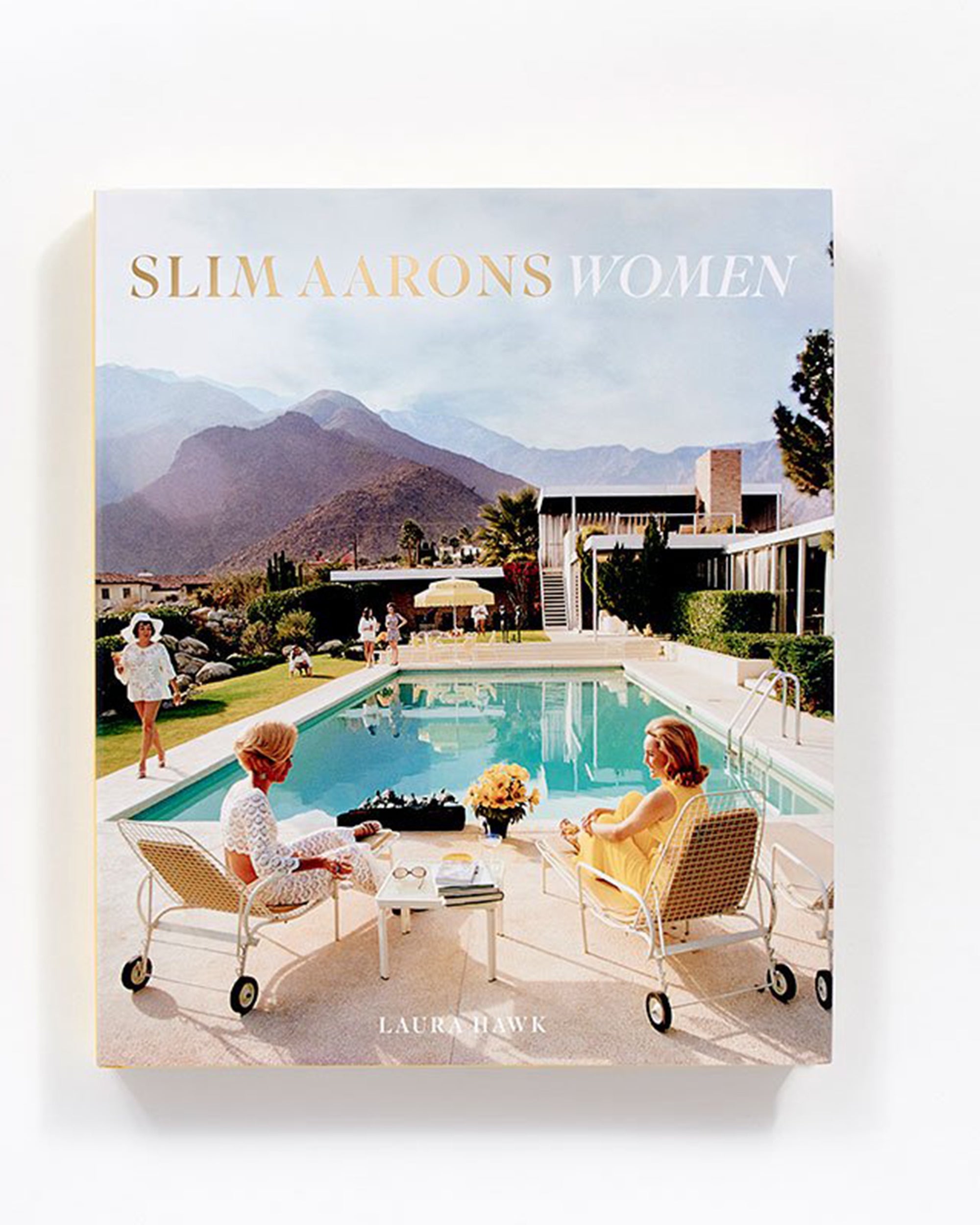The cover of "Slim Aarons: Women" by Hachette Book Group features elegant women in white outfits lounging poolside, with a modern house and mountains reflecting the high society glamour of the iconic photographer's work.