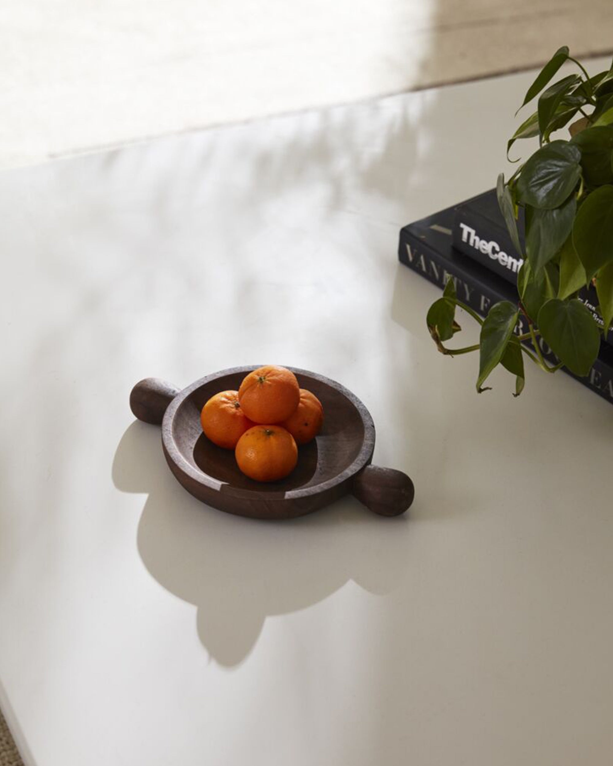 The Accent Decor Brownwood Tray /Small holds small oranges on a white table beside books and a green plant in sunlight, making it perfect for stylish coffee table decor.