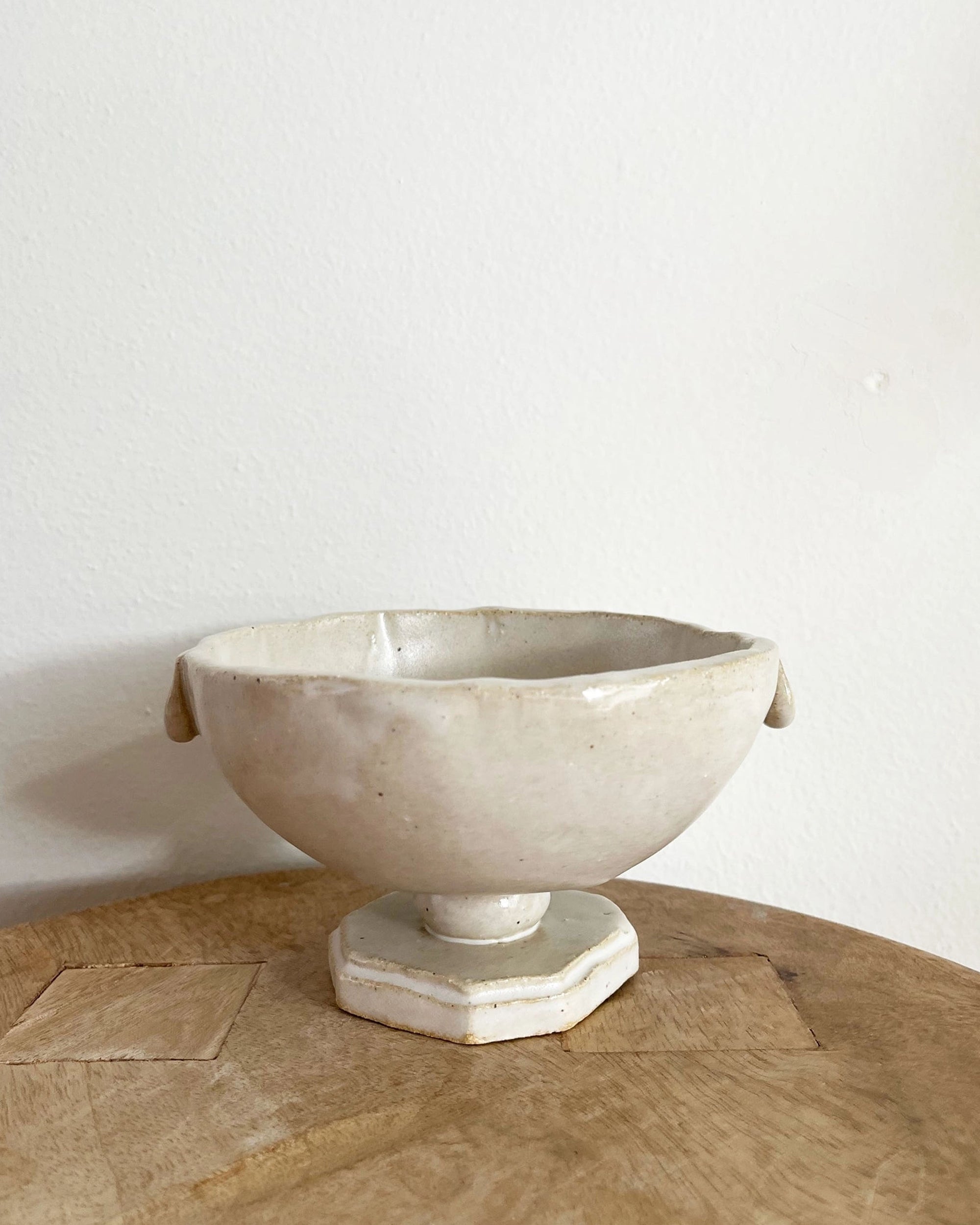 The Faire Tiny Trophy Bowl is a handmade off-white stoneware dish with a textured finish, irregular rim, and octagonal base, making it a unique trinket dish. Displayed on a light wooden surface against a plain white wall.