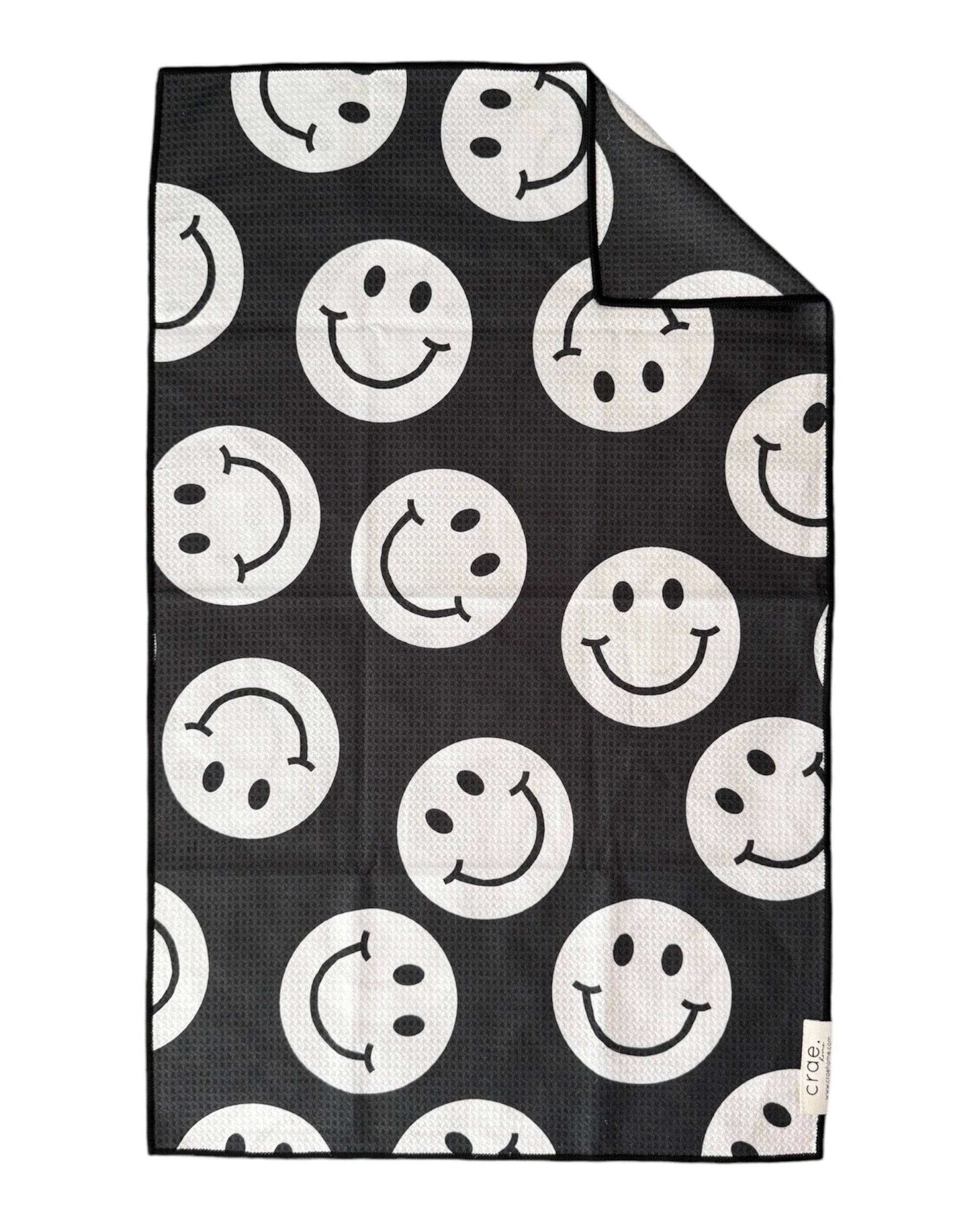 The Faire "Say Cheese" hand towel is black with a waffle weave and a pattern of large white smiley and frowning faces. A corner is folded to show the reverse side and an attached tag.