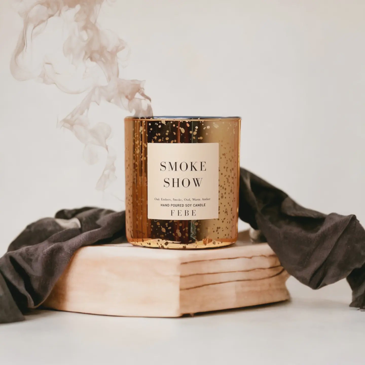 A Faire scented candle labeled &quot;Smoke Show&quot; on a book, surrounded by a gray scarf styled like an Arizona bungalow, with wisps of smoke rising from the FEBE candle, set against a soft off-white background.
