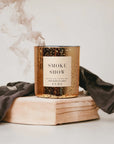 A Faire scented candle labeled "Smoke Show" on a book, surrounded by a gray scarf styled like an Arizona bungalow, with wisps of smoke rising from the FEBE candle, set against a soft off-white background.