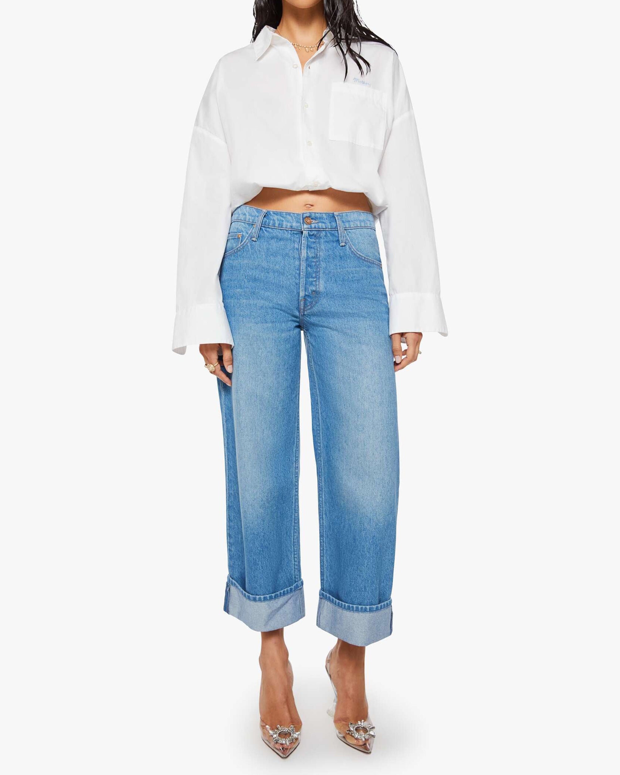 A person wearing Mother’s The Smoothie Crop Cuff jeans and a loose white button-up shirt with rolled-up sleeves, paired with clear pointed-toe heels, stands against a plain white background.