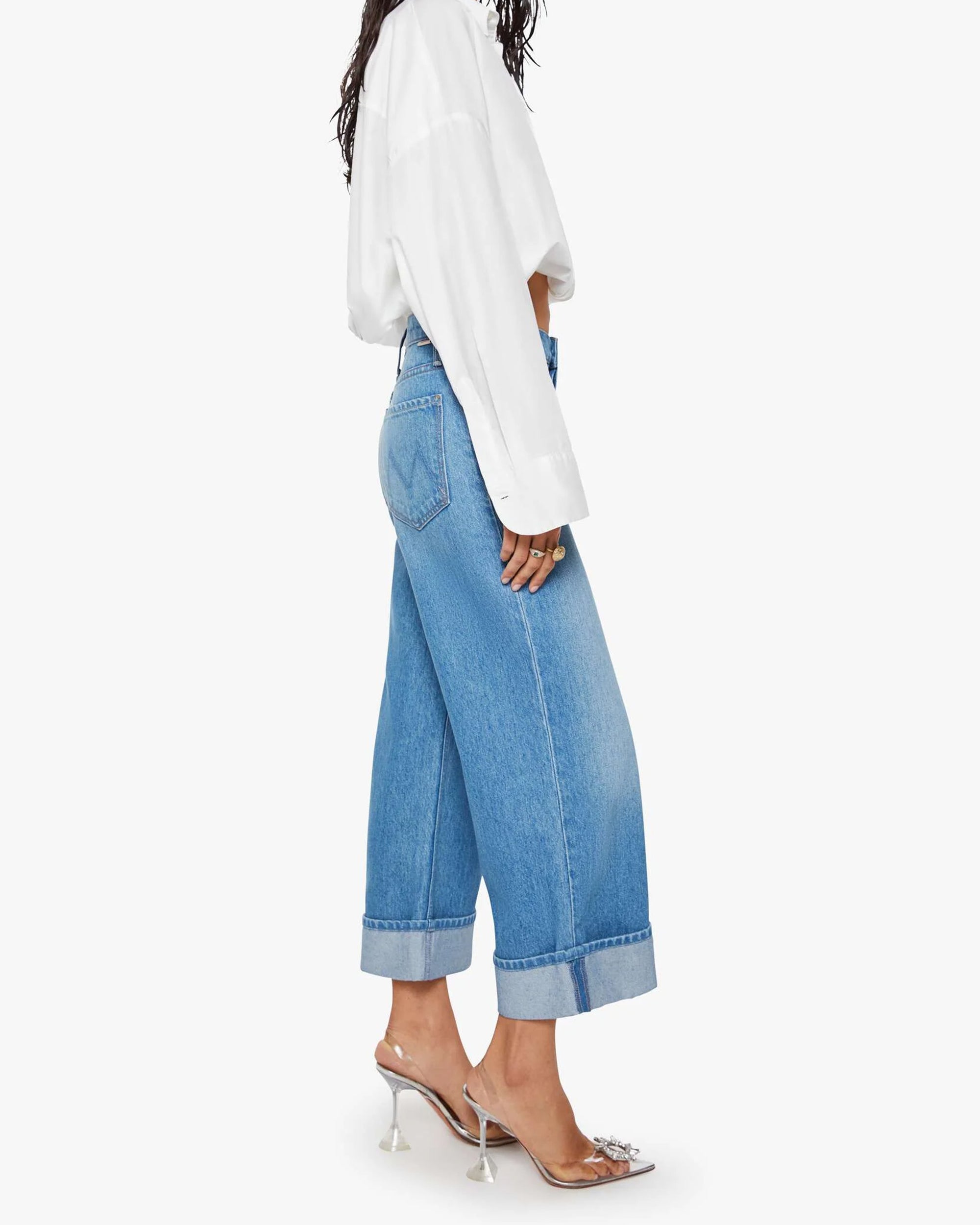 A person wearing Mother’s The Smoothie Crop Cuff high-waisted wide-leg jeans, a loose white shirt, and clear heeled mules stands in profile against a white background.