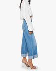A person wearing Mother’s The Smoothie Crop Cuff high-waisted wide-leg jeans, a loose white shirt, and clear heeled mules stands in profile against a white background.