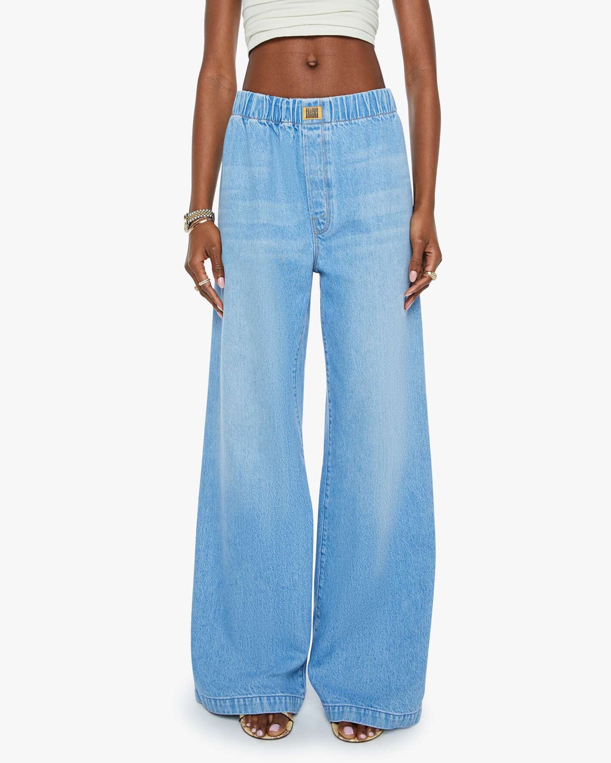 Someone wears The Kitchen Drawer Skimp by Mother—light blue, high-rise wide leg jeans crafted from a recycled cotton blend with an elastic waistband and pockets—paired with a cropped white top, posed against a plain background.