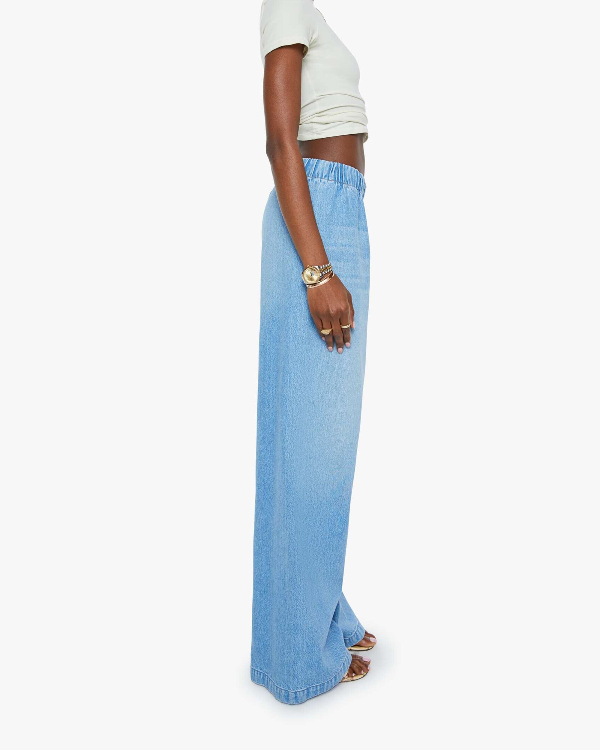 A person stands sideways against a white background, wearing Mother’s The Kitchen Drawer Skimp high-rise wide leg jeans in a recycled cotton blend with an elastic waistband, paired with a white cropped top, sandals, and a gold wristwatch.