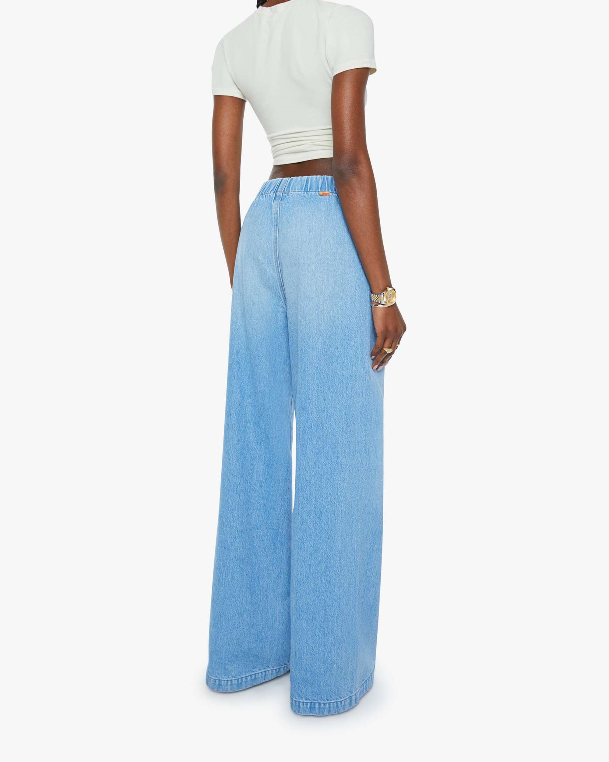 A person wearing Mother’s The Kitchen Drawer Skimp—a pair of high-rise, wide-leg jeans crafted from a recycled cotton blend—stands with their back to the camera, styled with a fitted white short-sleeve top against a plain white background.
