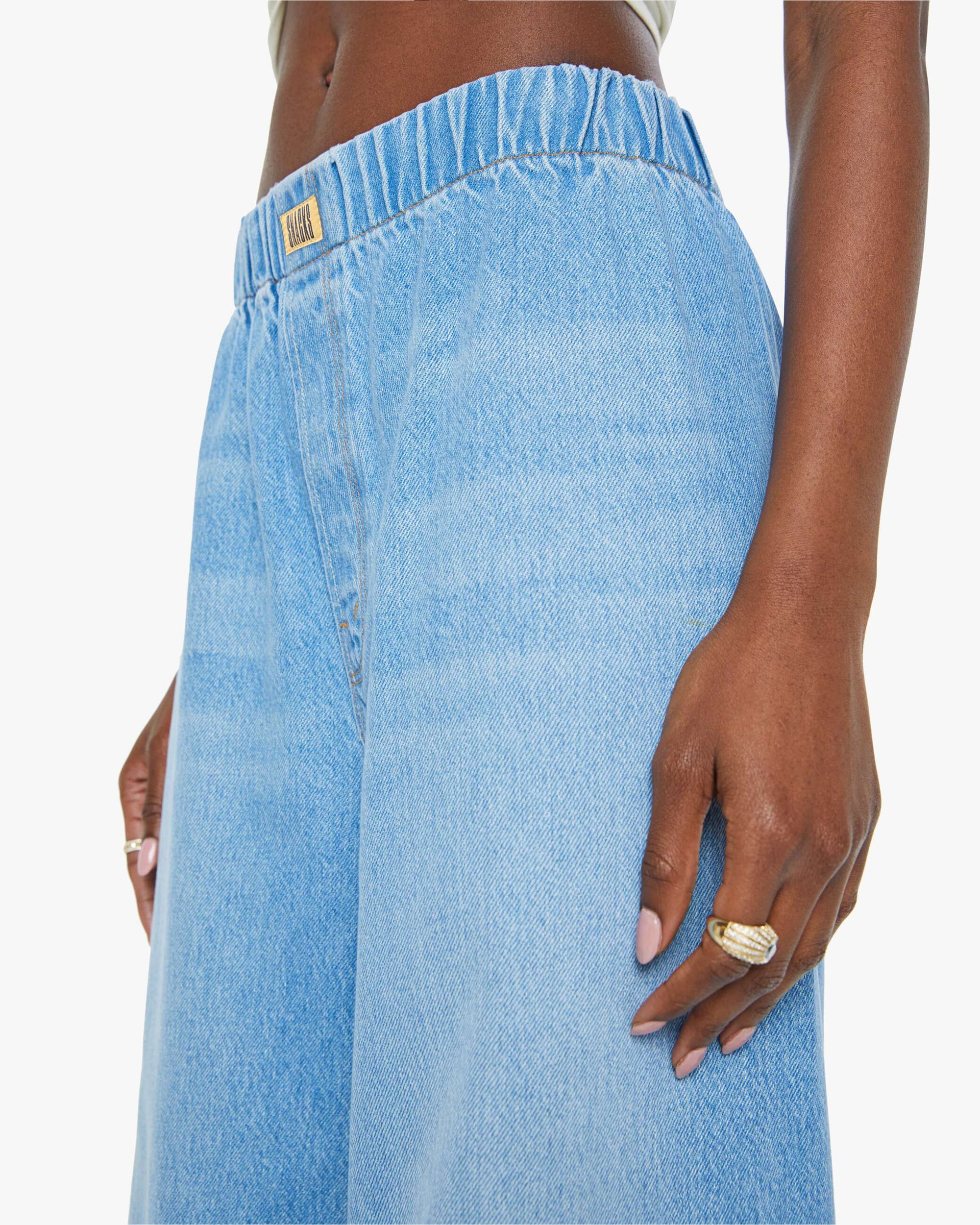 Close-up of a person wearing Mother’s The Kitchen Drawer Skimp light blue wide leg jeans with a high-rise elastic waistband in recycled cotton blend. Gold ring, light pink manicure. Plain white background.