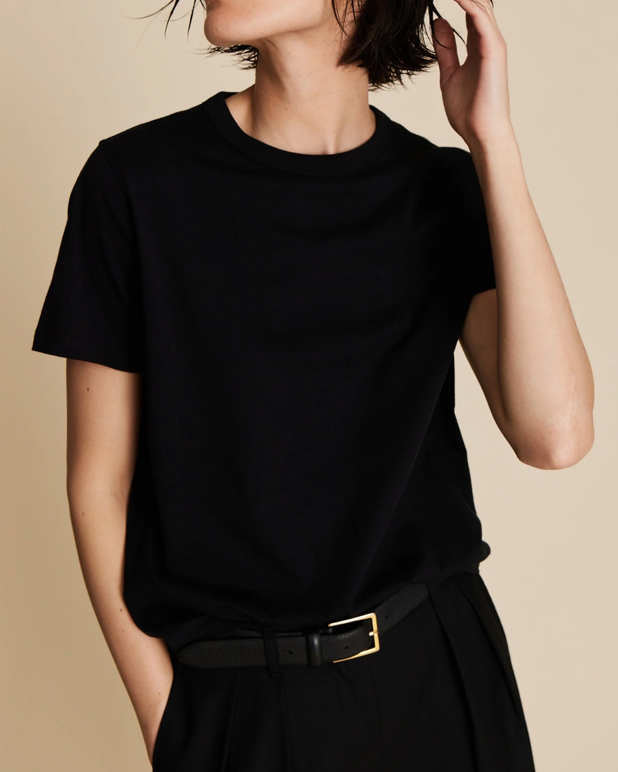 Wearing The Soft Perfect Tee by Sold Out NYC, this person stands against a neutral background in black pants and a belt, one hand in their pocket and the other touching their hair, with their face partially out of frame.