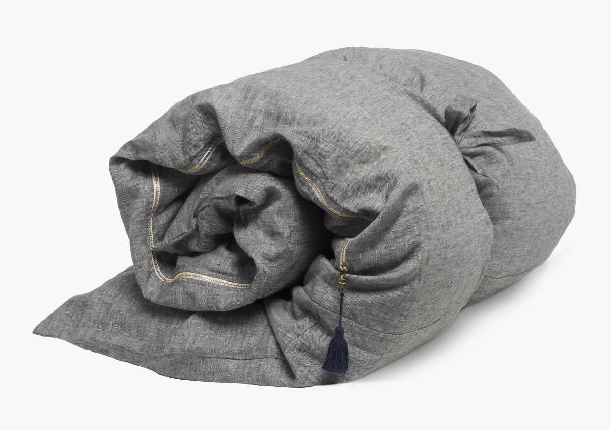 The Hedgehouse Throwbed is a rolled-up gray duvet with yellow stitching, a small dark blue tassel on one corner, and cotton-linen blend covers, shown on a plain white background.