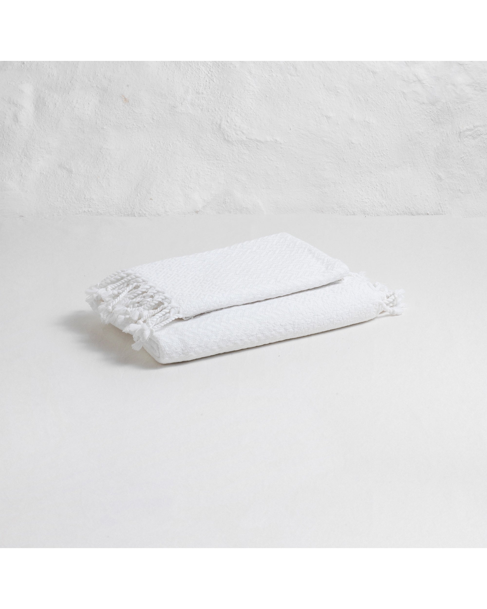 The Faire Spa Hand Towel, hand-loomed in white with tasseled edges, is folded on a light surface against a textured white wall.